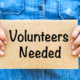 Person holding a sign reading volunteers needed